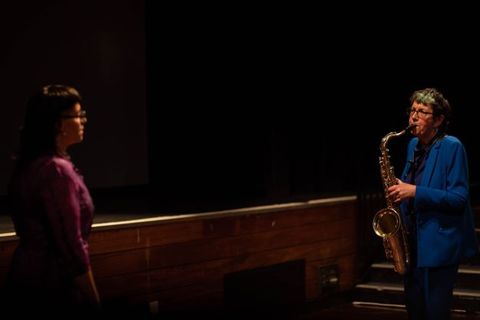 Performance art. Two women stand facing each other, the left woman wearing glasses and a fitted pink silk dress. A saxophone-playing woman with short hair and a mid-blue suit looks towards the first woman in front of a stage and steps in this colour photograph.