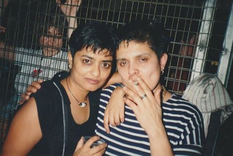 Colour photograph. Two people from the shoulders up face the camera with their arms around one another. One is smoking a cigarette while the other holds a partially seen plastic cup. Behind them is a metal barricade, through which we can see another person in the background.