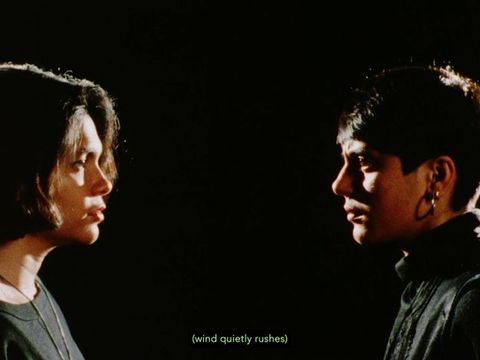 Colour film still. Two women stand at either side, looking into each other's brown eyes. One has a bob hairstyle, grey top and metallic gold necklace. The other wears shiny gold hoop earrings, short hair with a fringe and embellished grey polo neck jumper. Green subtitle text "wind quietly rushes" against a black background.