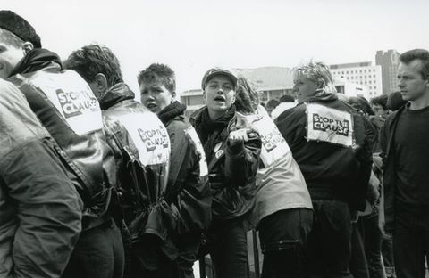Black and white photograph. Group of six people wearing dark wax jackets featuring the "STOP THE CLAUSE" capitalised text and protest symbol; an inverted triangle intersecting the text. A seventh person looks leftwards towards the group, the majority of whom have their backs turned.