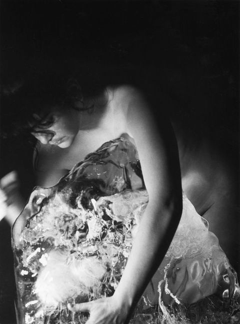 Black and white photograph. Naked woman with curly fronds of hair across her forehead curves her back and leans towards the left, her right hand is blurred while her left hand is placed on a partially jagged semi-transparent object. Solid black background.