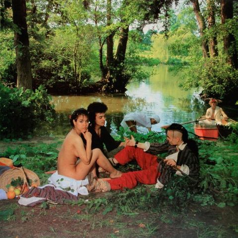 Colour photograph. Three people are seated on the ground beside a lake, surrounded by trees. Two are fully clothed and hold hands. The third is nude, with arm and leg bent to partially conceal their body, while a picnic basket spills out food beside them. In the distance a partially dressed person leans forward to wash themselves in the lake, while another person sits in a small rowing boat. 