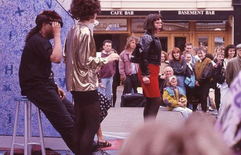 Performance art. A woman wearing a loose metallic blouse standing by a guy dressed in black clothes sitting on a stool. Another woman in the middle ground has a bob, leather jacket, fitted red mini skirt and glasses, her mouth opened. The on-street crowd laughs in the background of this colour photograph.