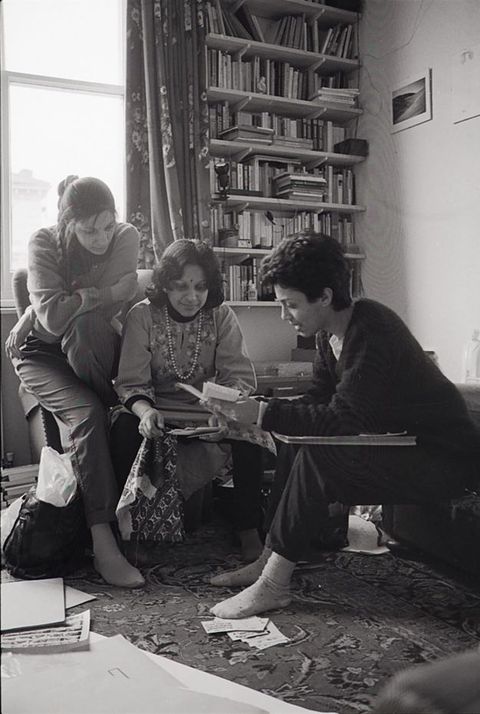 Black and white photograph. Three South Asian women look at postcards in the centre of a room. The middle, older woman sits on an armchair while a younger woman with plaited hair sits on the chair’s arm, her right leg touching an oriental-style carpet. A short-haired woman leans towards the others, her left foot covering postcards on the floor.