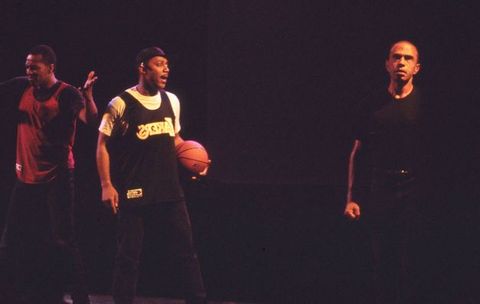 Performance art. Two men with dark skin tones standing to the left wearing basketball jerseys, one holding his arms at 90 degrees, the other a textured orange ball in left hand. Another (light skin tone) man stands half in shadow, clenched hand, wearing a black t-shirt and trousers in this onstage colour photograph.