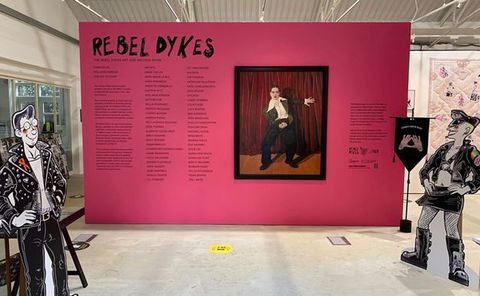 Installation photograph. A freestanding pink wall is emblazoned with text in a hand-drawn style reading ‘REBEL DYKES”. Below the text is a longer passage of text, accompanied by an artwork featuring a person dressed in a black suit against a red curtain. Two freestanding, life-size cut-out illustrations of people dressed in black leather are posed in the space.