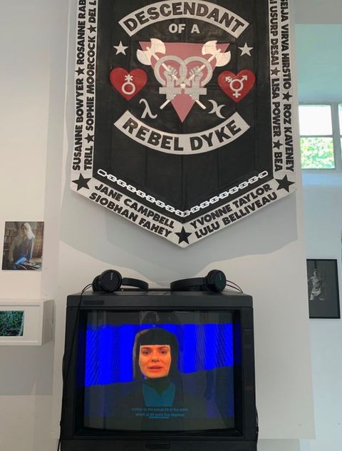 Installation photograph. A black, white, red and pink banner featuring the words “DESCENDANT OF A REBEL DYKE” and a heraldic logo is hung on a white gallery wall, over a television monitor on which a person’s head and shoulders are visible against a blue curtain. 