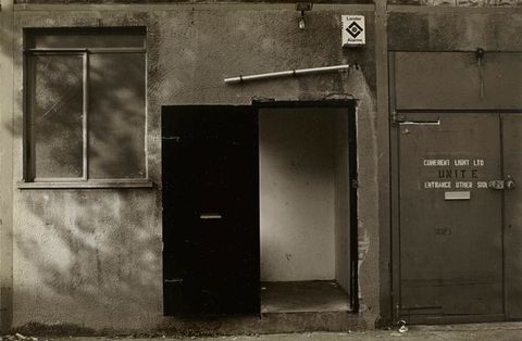 Black and white photograph. Close-up of a rendered concrete building and black door with a letterbox is open in the centre, near a window and security alarm box. Capitalised stencilled text reads "ENTRANCE OTHER SIDE".