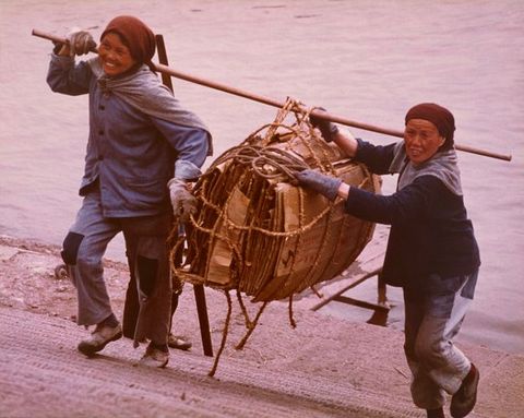 Colour photograph. Two Chinese women in working clothes and headscarves stand carrying a bamboo pole which holds folded brown cardboard boxes tied with rope. They smile while walking up the steps, appearing in front of a body of water in the background.