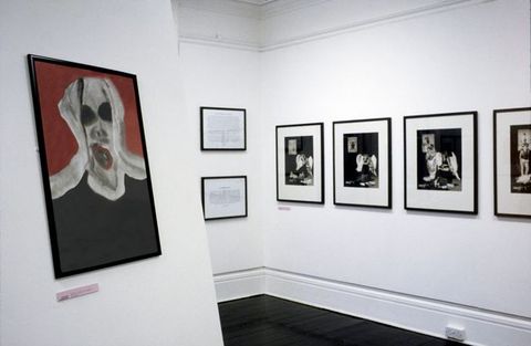 Installation photograph. A view into a gallery space with white walls and dark floors. On the right, a series of black and white, framed photographs are hung on the wall, and on the left is a red and black painting of a person’s head. 