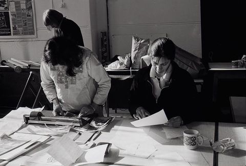 Black and white photograph. Three women work in an office or studio space. Two of them are looking down at a desk. One wears an Asian tunic, trimming photographic prints using a guillotine, while another on the right reads handwritten letters. Another woman stands working behind them.