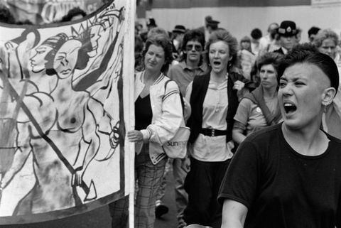 Black and white photograph. An open-mouthed woman stands looking past the left of the frame. A row in the middle ground includes three women protestors, the left woman holding a banner of three muscular naked figures, scythes and a spear. A uniformed policewoman stands in the background, looking towards the foreground.