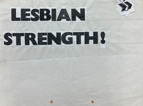 Black and white photograph. This photograph documents the mock up for a flyer, with cut-out, individual black and white letters pasted on a white sheet of paper reading, “LESBIAN STRENGTH”. A stack of further cut-out symbols is attached to the paper on one corner by a paperclip.