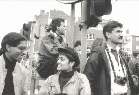 Black and white photograph. Four people standing in a rhombus arrangement at a traffic light. A central woman wearing a cloth hat and denim jacket talks to a man wearing glasses on the left. Another man stands to the right of her, a camera around his neck. The background street has apartments and shops.
