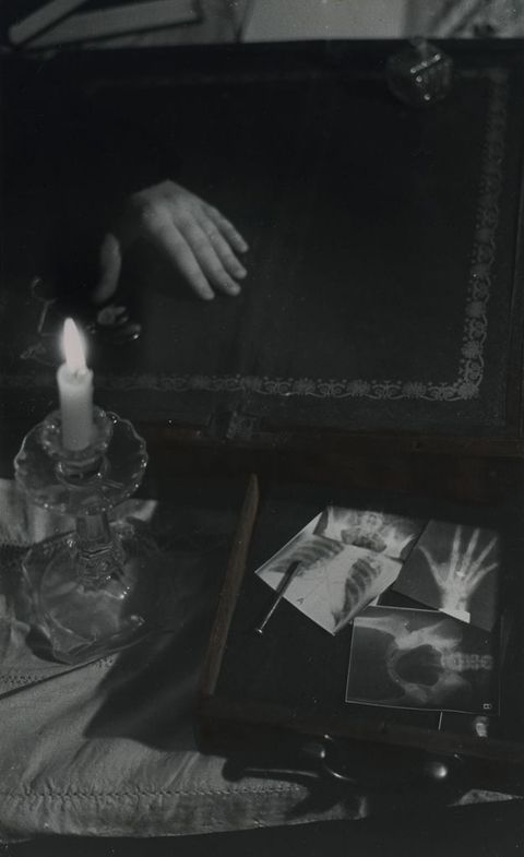 Black and white photograph. A dark interior scene is lit by a small candle in a glass holder on the left, which rests on a flat chair cushion. On the right, the interior of a pulled-out desk drawer contains x-rays of a hand, a ribcage, and a pelvis. Beyond them a human hand emerges out of the darkness, resting palm down on an antique, fold-out table top.