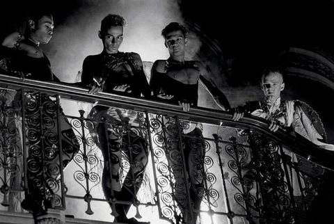 Black and white film still. Three men wearing matching black studded leather harnesses and draped shorts stand on a wrought iron balcony with curled metalwork, a fourth man on the right wears feathered wings and a cloth tunic. Clouds of smoke and ornate arches fill the background.