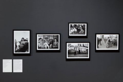 Installation photograph. Black and white. Five photographs in dark frames with light paper mounts are displayed on a grey wall. From left-to-right, the photographs depict an Indian person seated holding a pipe, an Indian family, an Indian dancer, two policemen walking past a store, and protestors outside a building holding an "Anti Facist Action" banner next to standing man and woman in a wheelchair.