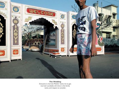 Colour photograph. Man with medium-brown skin wearing pastel pink and green denim shorts and t-shirt stands in front of an ornate wooden gateway decorated in gold and bright colours. Typewritten text including ""The Wedding"" spans the lower quarter of the page on a white background."