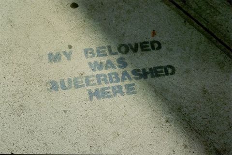 Colour photograph. Textured concrete floor half-covered in shadow and sunlight, with the capitalised grey stencilled text "MY BELOVED WAS QUEER BASHED HERE". The floor has mottled textures and two parallel lines seen in the top-right corner.