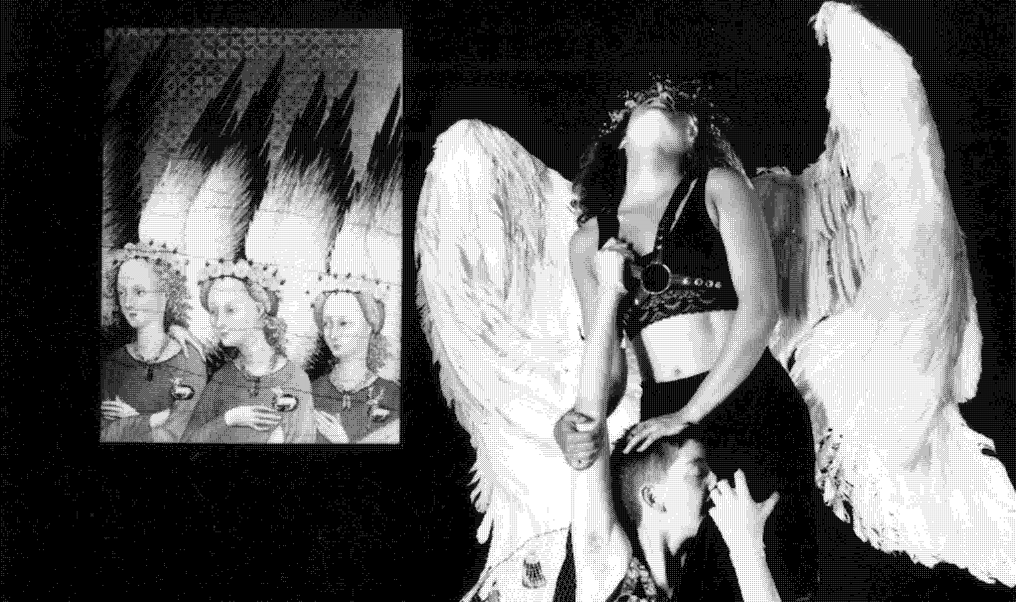 Detail of a black and white photograph. A woman wearing large, white angel wings sits intimately on the shoulders of another, who is wearing mesh wings adorned with metal objects. Magazines, a sex toy and a toy alligator are strewn on the floor below, while an artwork depicting three angels is projected onto the wall. 
