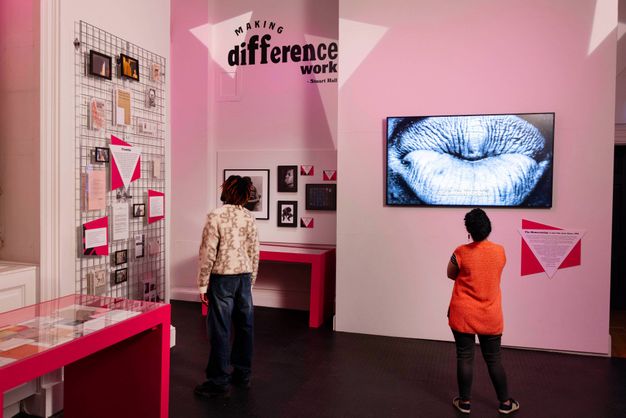 Installation. A man and woman with dark-brown skin tones stand viewing various artworks including black and white photograph of lips, and reoccurring white inverted triangle against a trapezoid pink shape. Corner view of pink room featuring the Stuart Hall quote "Making difference work" in black lettering.